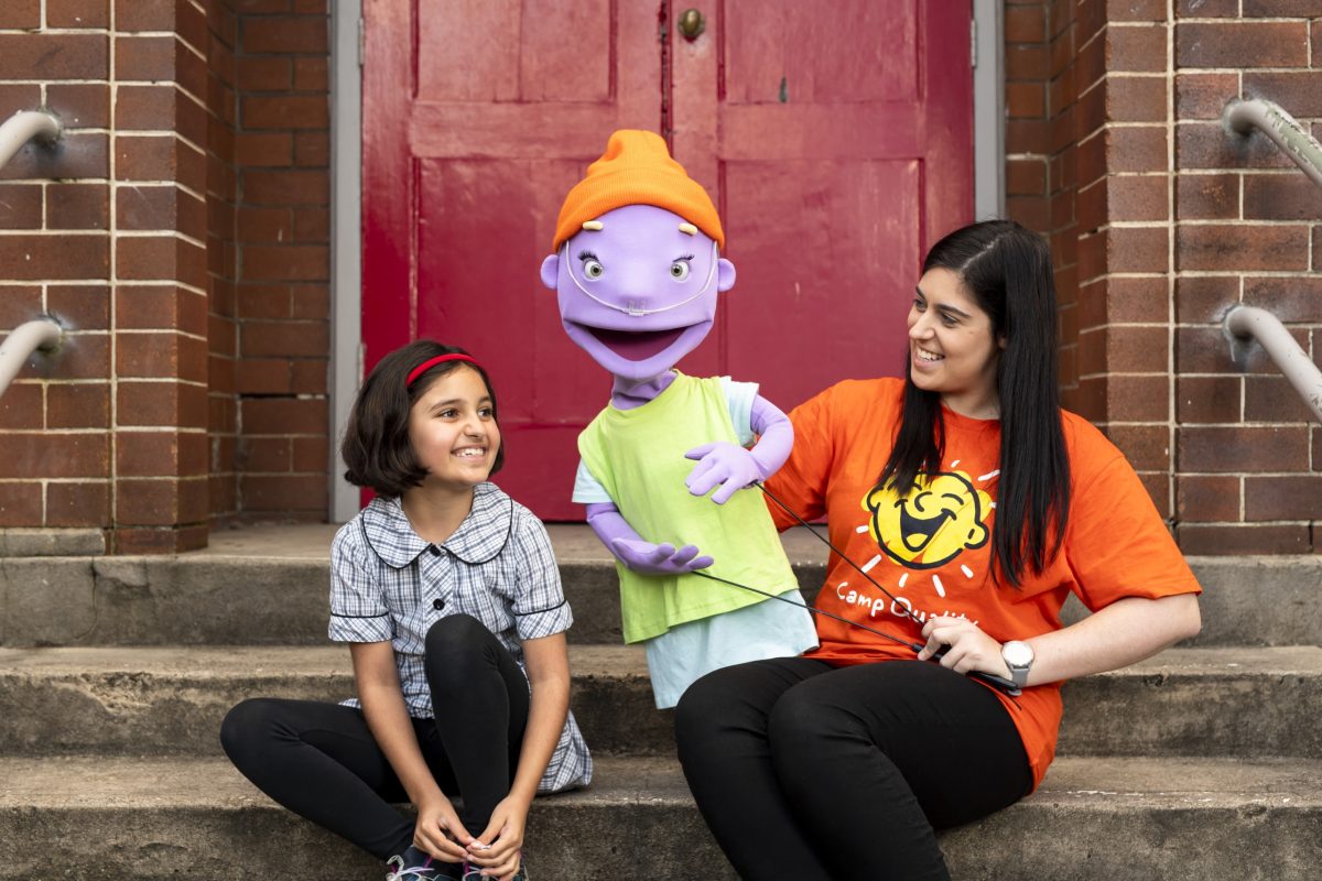 Lady using a camp quality puppet, sitting next to a school girl on a set of stairs
