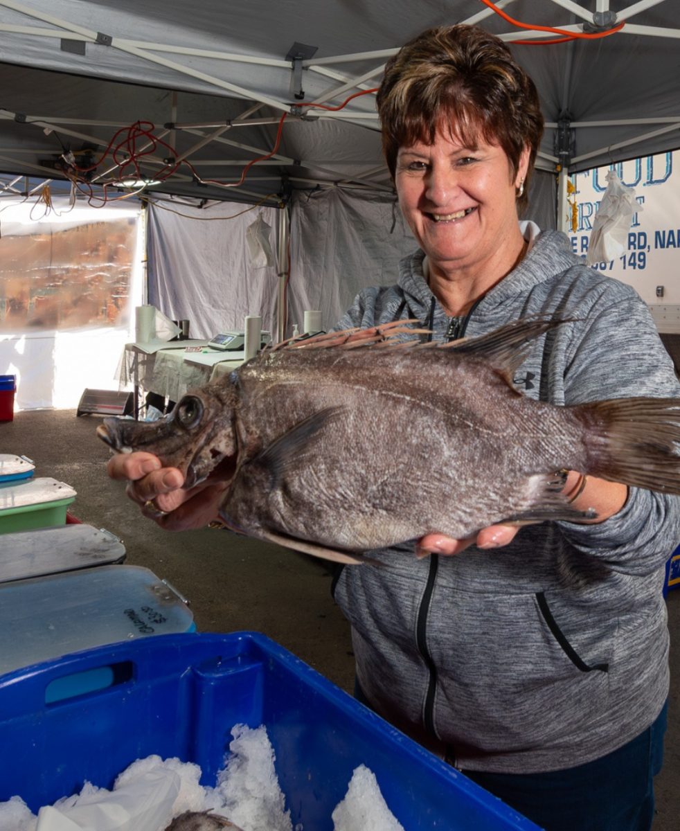 Woman smiles and holds a large fish at a market stall.
