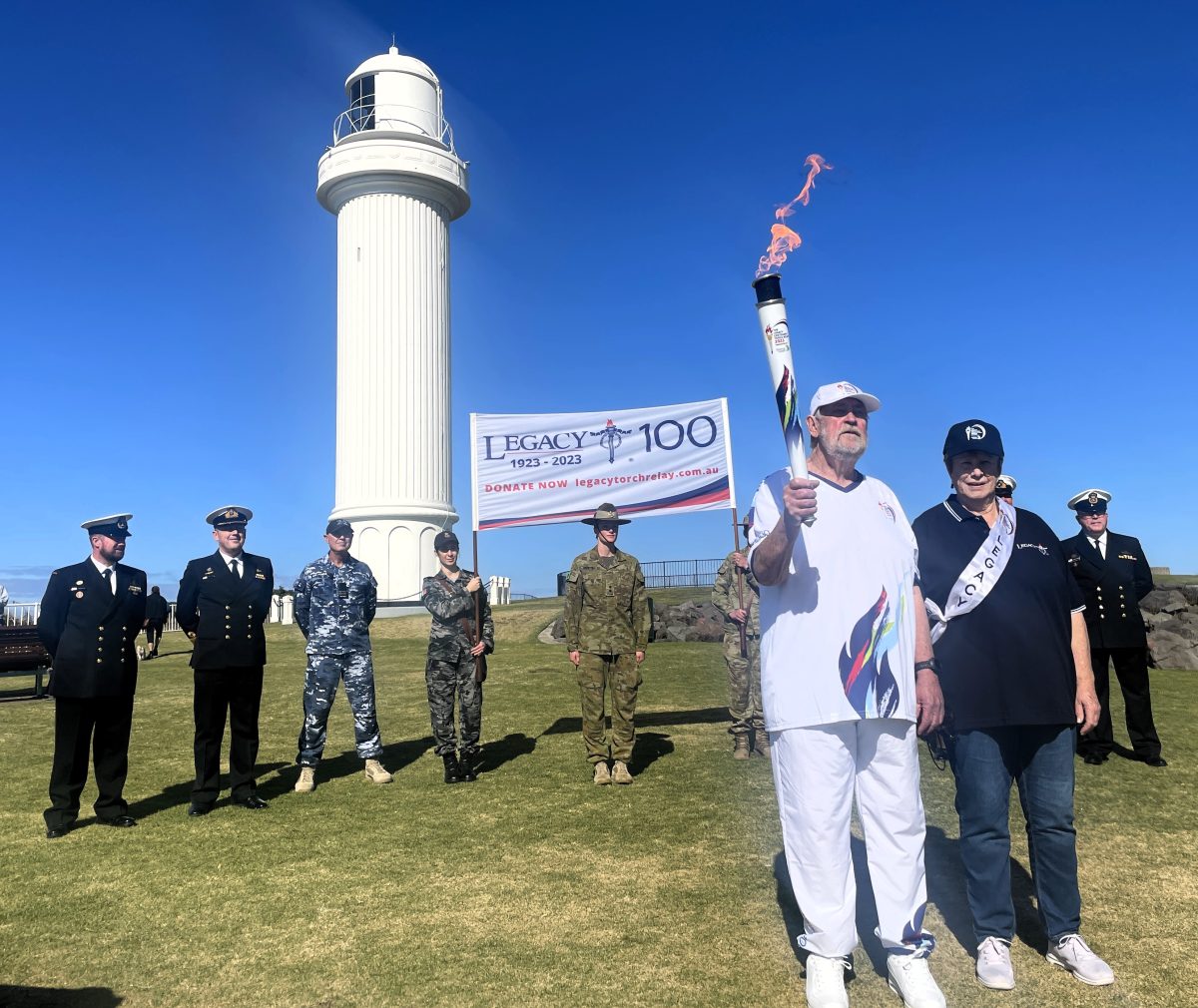 Greg Keir holding torch with servicepeople and Legacy sign behind him at Wollongong Lighthouse.