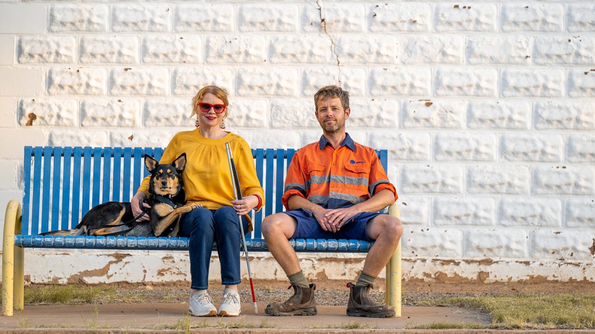A blind woman, her dog and a tradie sit on a bench