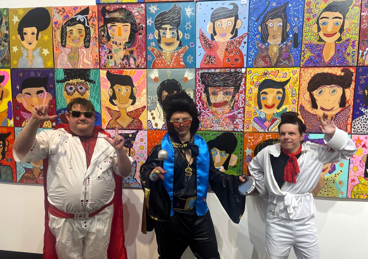Three artists dressed as Elvis in front of Elvis portraits and gallery exhibition. 