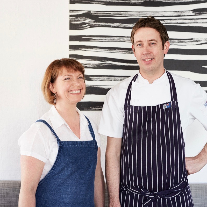 Sonia smiles in a blue apron and John has one hand on his hip in a striped apron. A piece of black and white art hangs on the wall behind them.