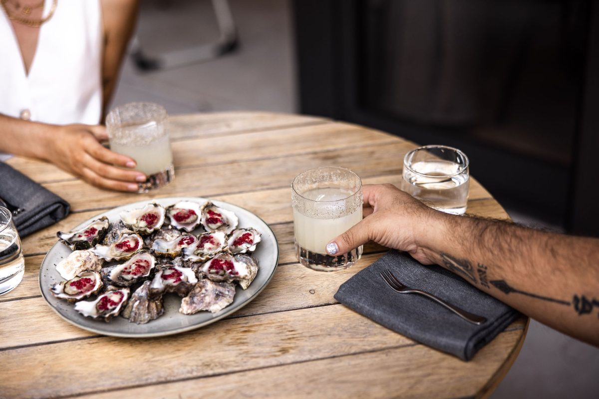 Plate of oysters with a pink garnish, two people hold drinks.