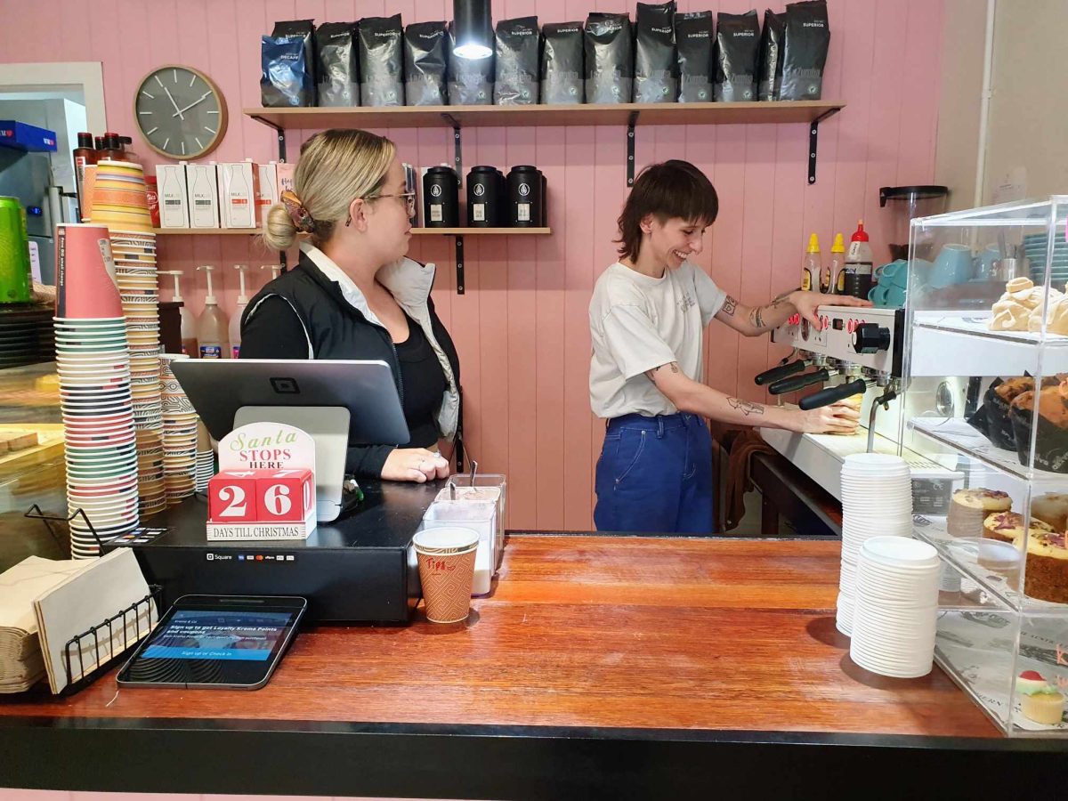 Front of house staff Asta Dundas and Chloe Suckalis prepare to serve customers and make coffee behind a wooden counter in front of a pink wall covered with coffee.