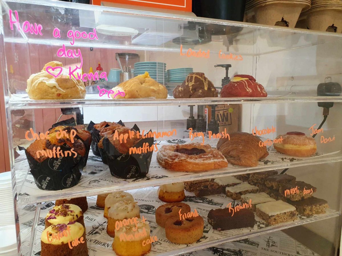 Cookies, muffins, cupcakes and slices are on display in a cabinet with neon-orange labels. 'Have a good day, Krema xxx' is hand-written on the cabinet in pink.