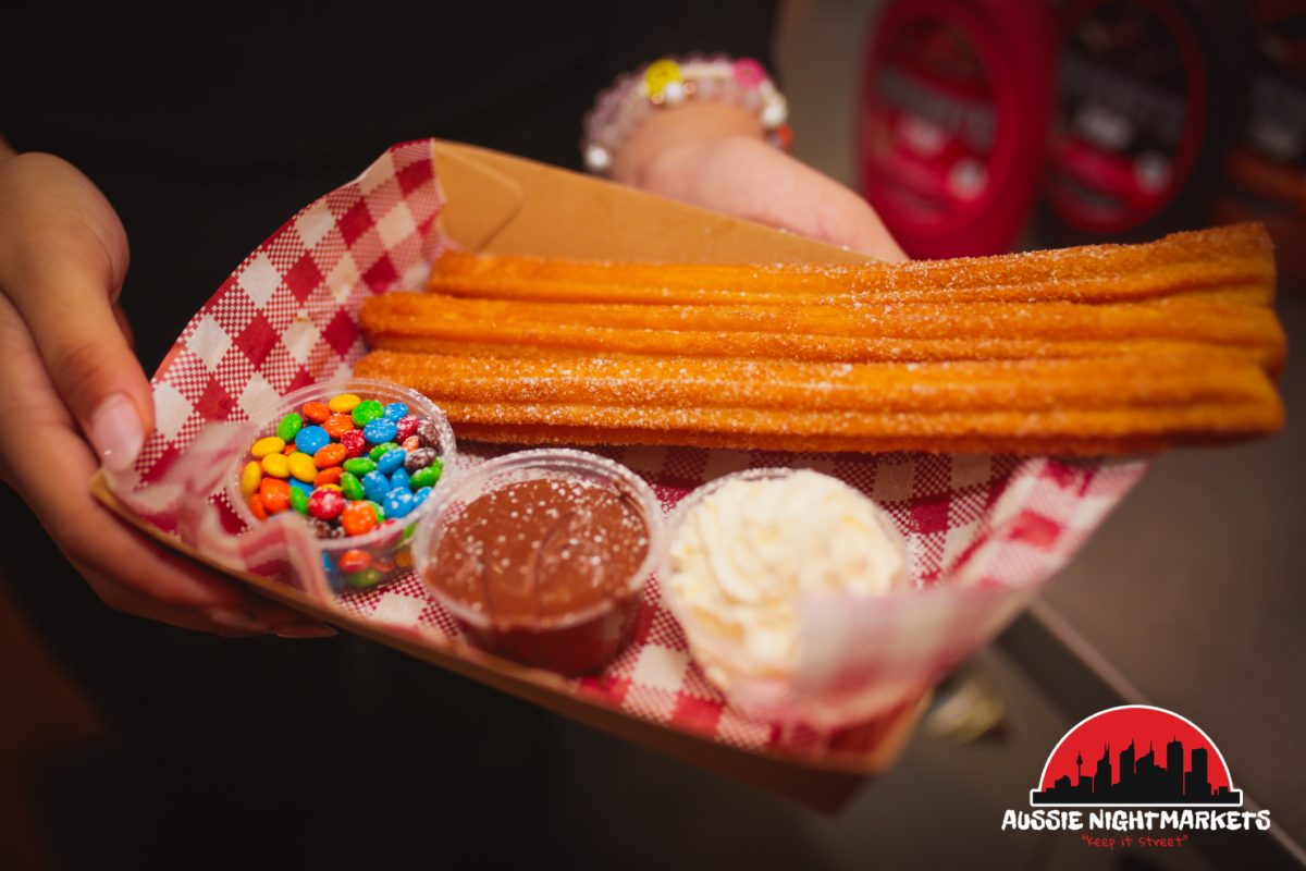 Churros with chocolate dipping sauce and brightly coloured sprinkles are presented on a cardboard takeaway tray.