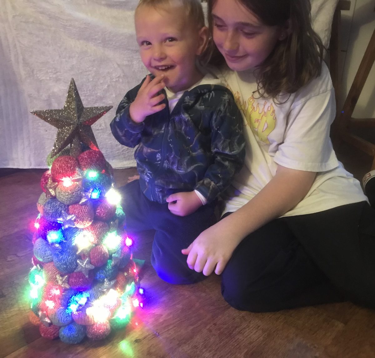 Two boys looking at a knitted, lit-up Christmas tree.