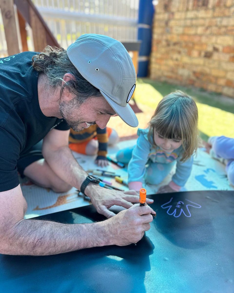 A smiling man helps a small child draw on a surfboard