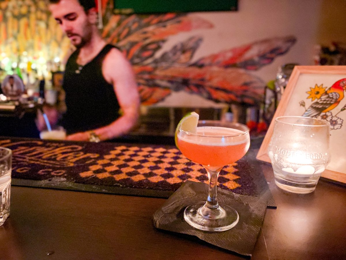 Pink cocktail in a coupe glass with bartender and mural in the background.