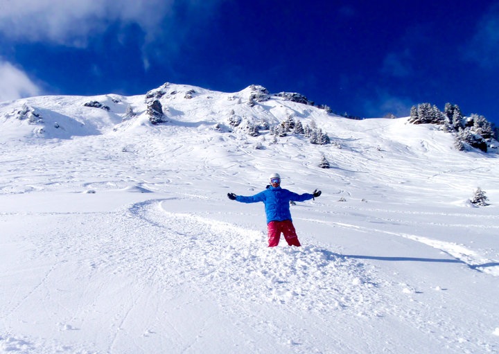 A man on a snowboard stands near the peak of a snow-covered mountain, his arms spread wide.