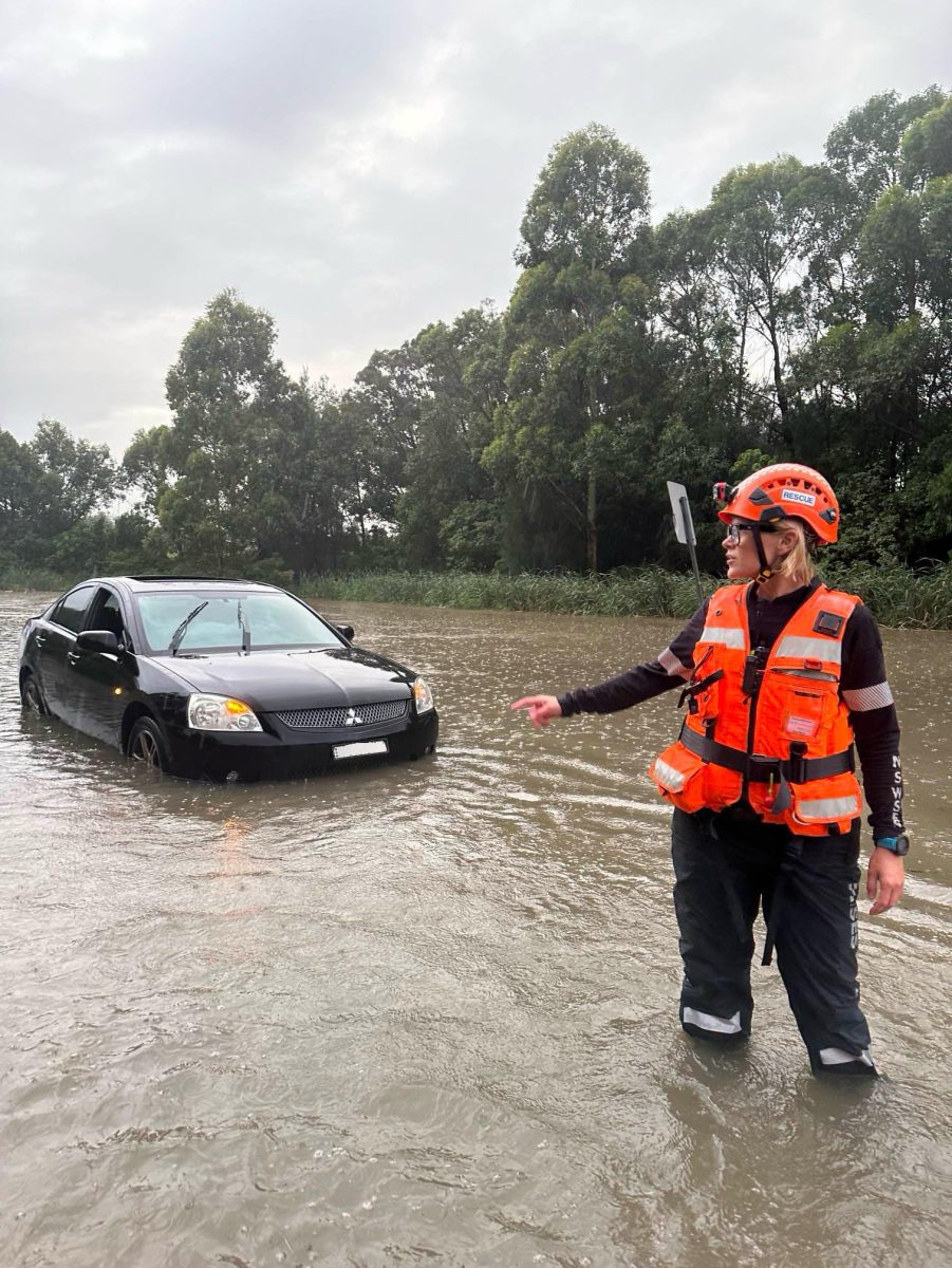 A woman in an orange hi-vis vest and hard hat stands in knee-high water with her arm out. She is looking at a car stuck in the water
