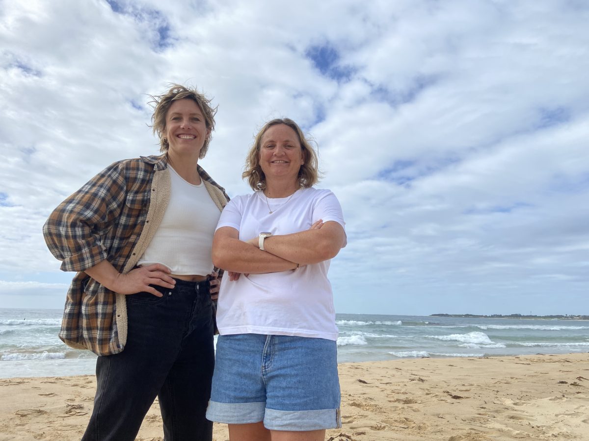 Anna Suthers of Reub Goldberg Brewing Machine stands on the beach with Yvonne Turner of Woonona Boardriders