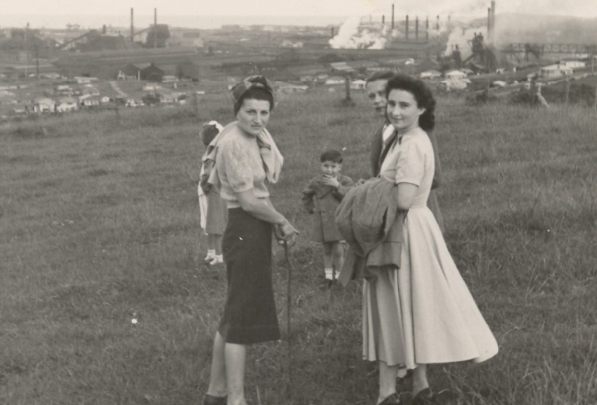 old photo of people walking through parkland, with steelworks in background