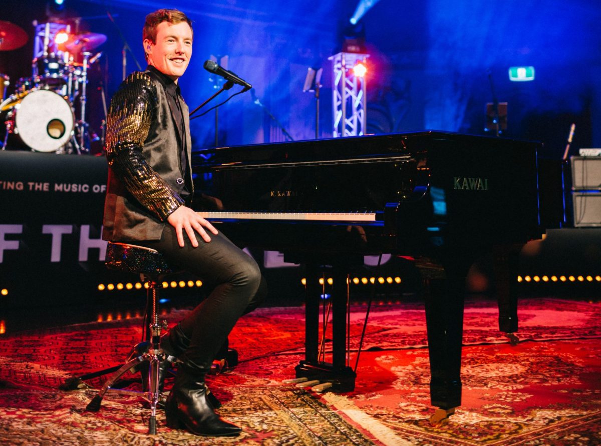 A smiling Liam Cooper sits at a piano on stage