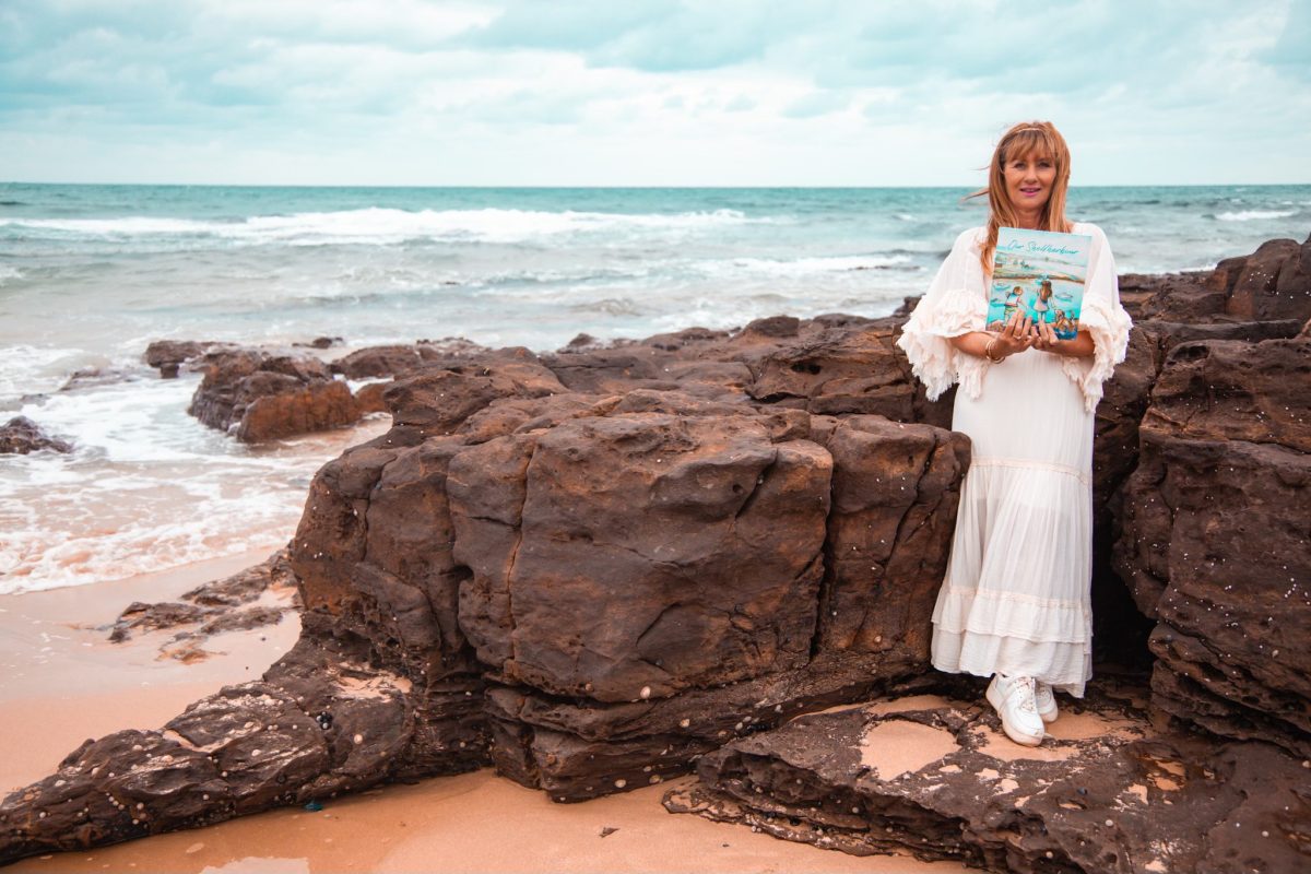 Author Karen Hendriks standing on the beach holding her book Our Shellharbour 