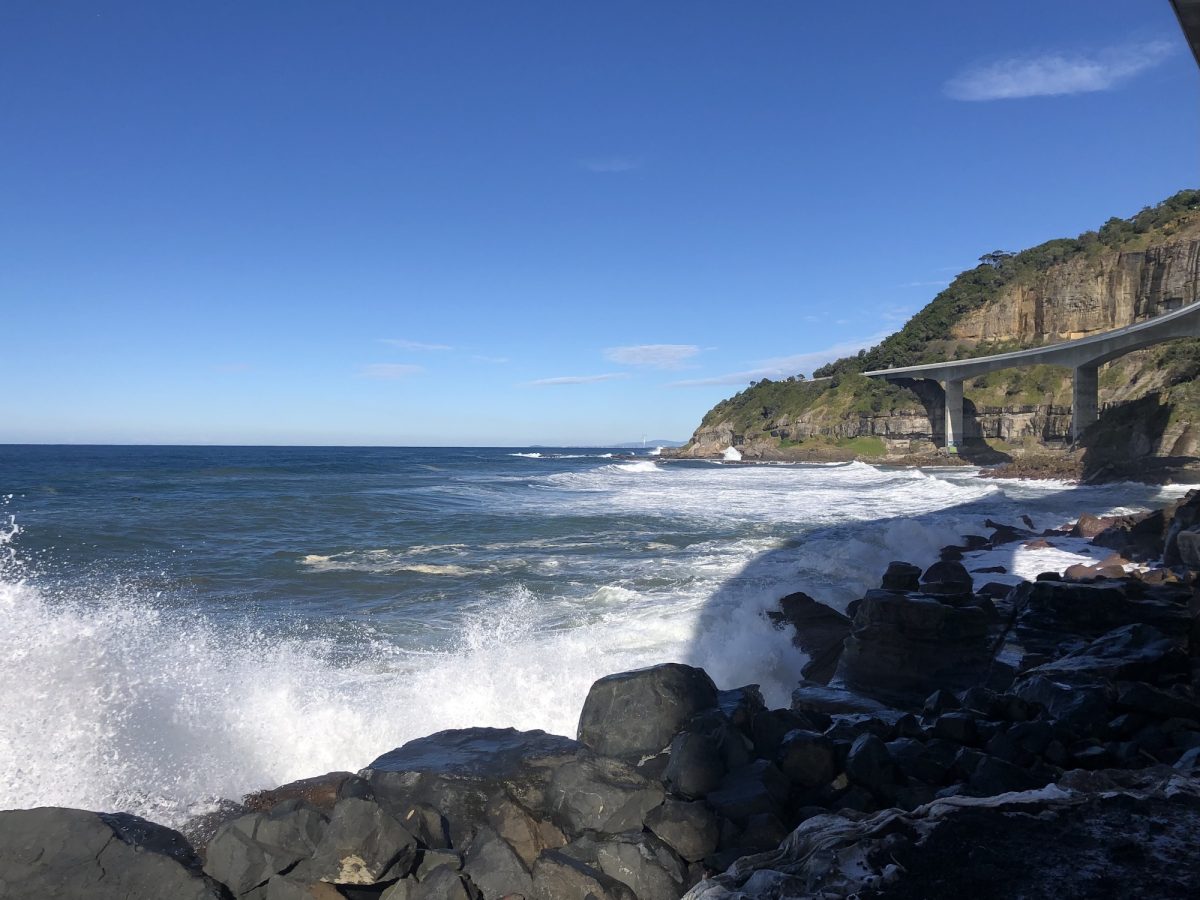 Waves crashing against rocks with Sea Cliff Bridge in the background.