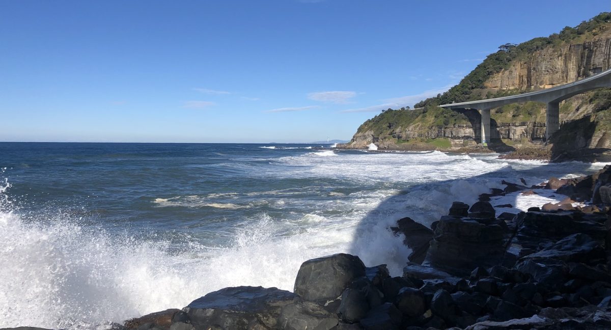 Waves crashing against rocks with Sea Cliff Bridge in the background.