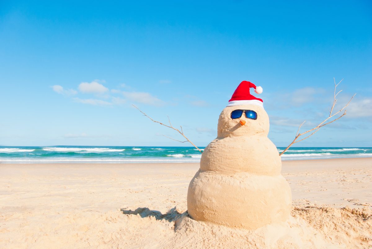 Snowman made of sand on the beach wearing a Santa hat and sunnies