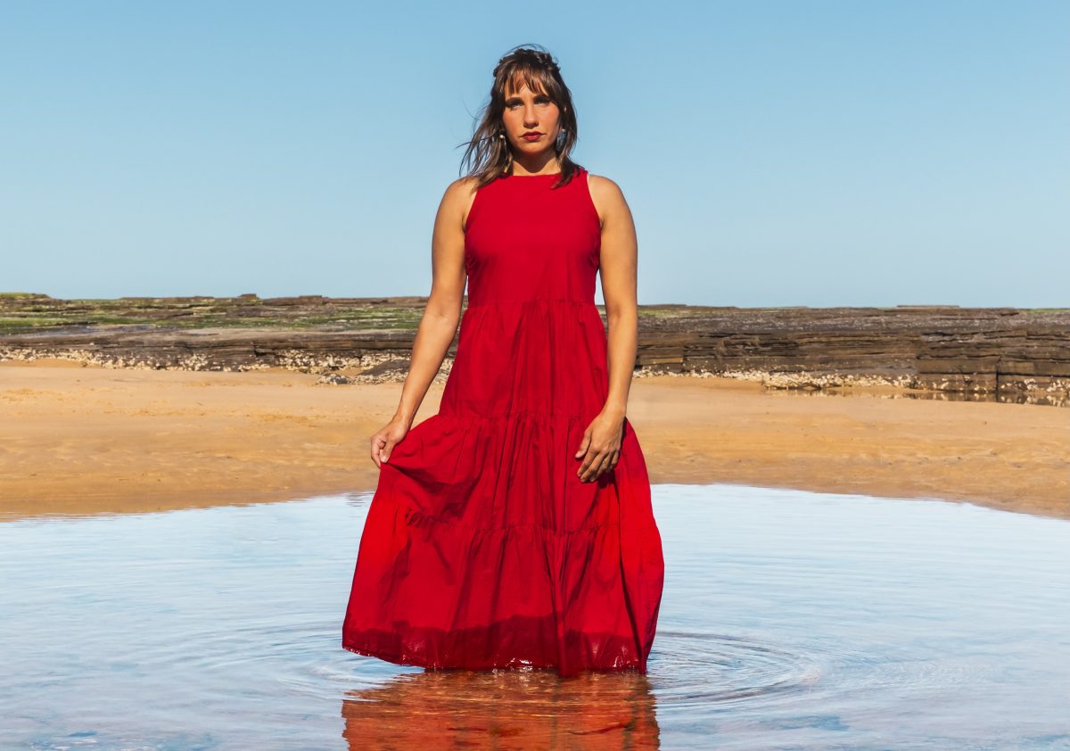 First Nations artist Kirli Saunders stands in water wearing a striking red dress.