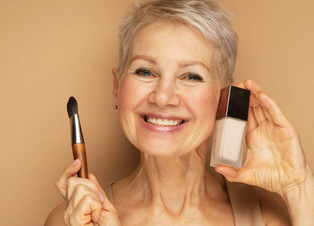 Charming elderly woman holds makeup brushes and foundation in her hands