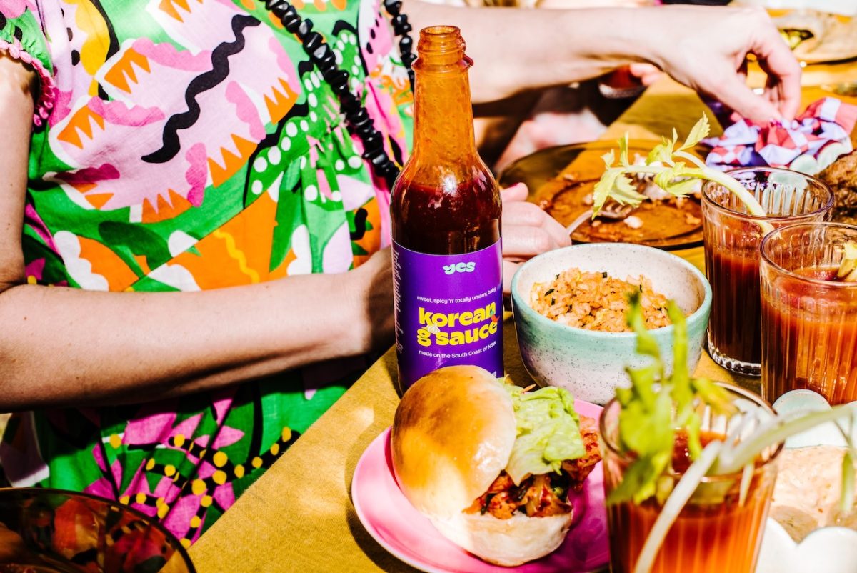 A busy photo of food, someone in a colourful dress and a bottle of sauce.