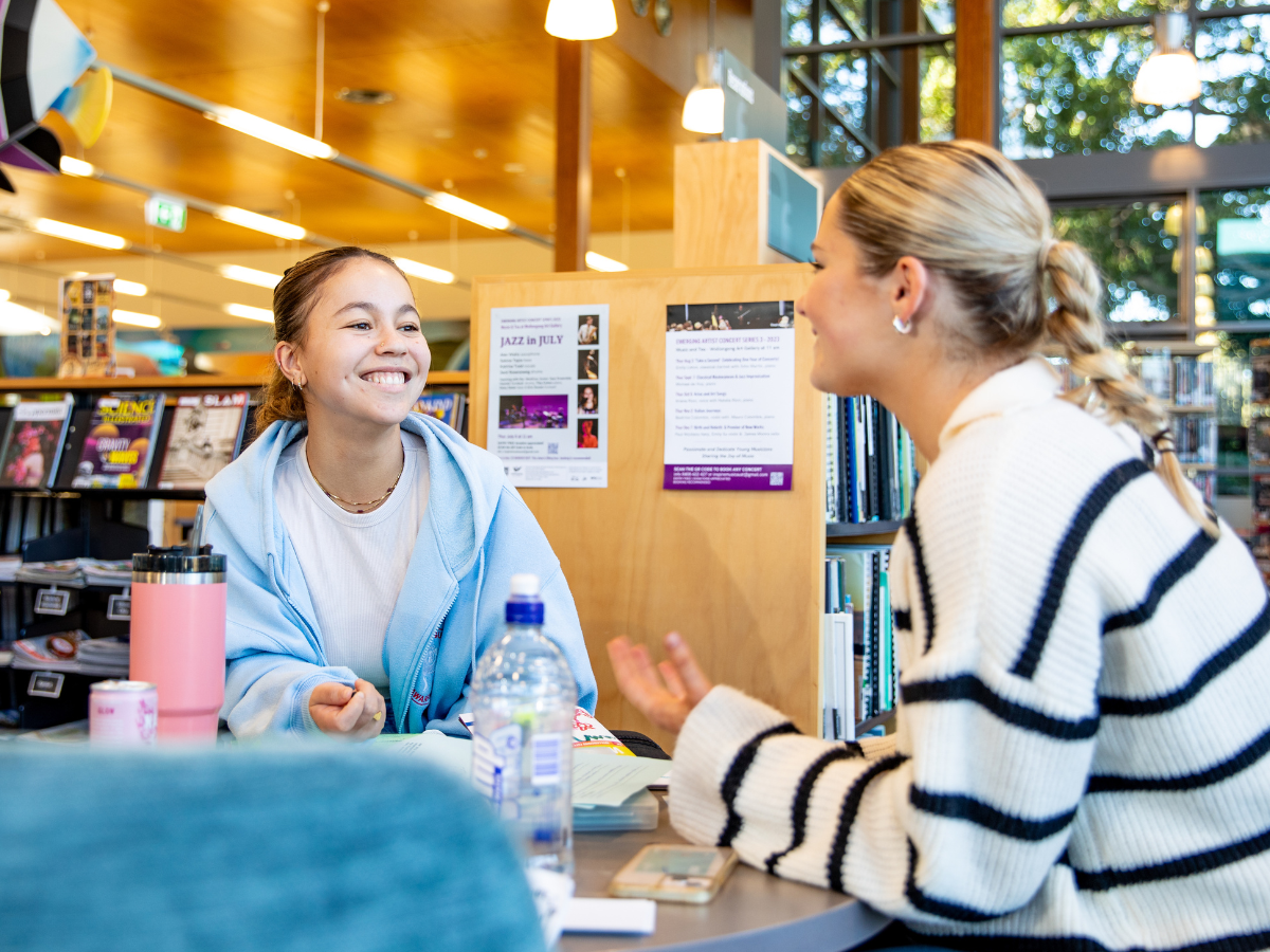 Two students sitting at a desk in a library talk to eachother