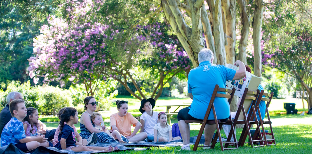 people sitting in a garden listening to someone reading a book