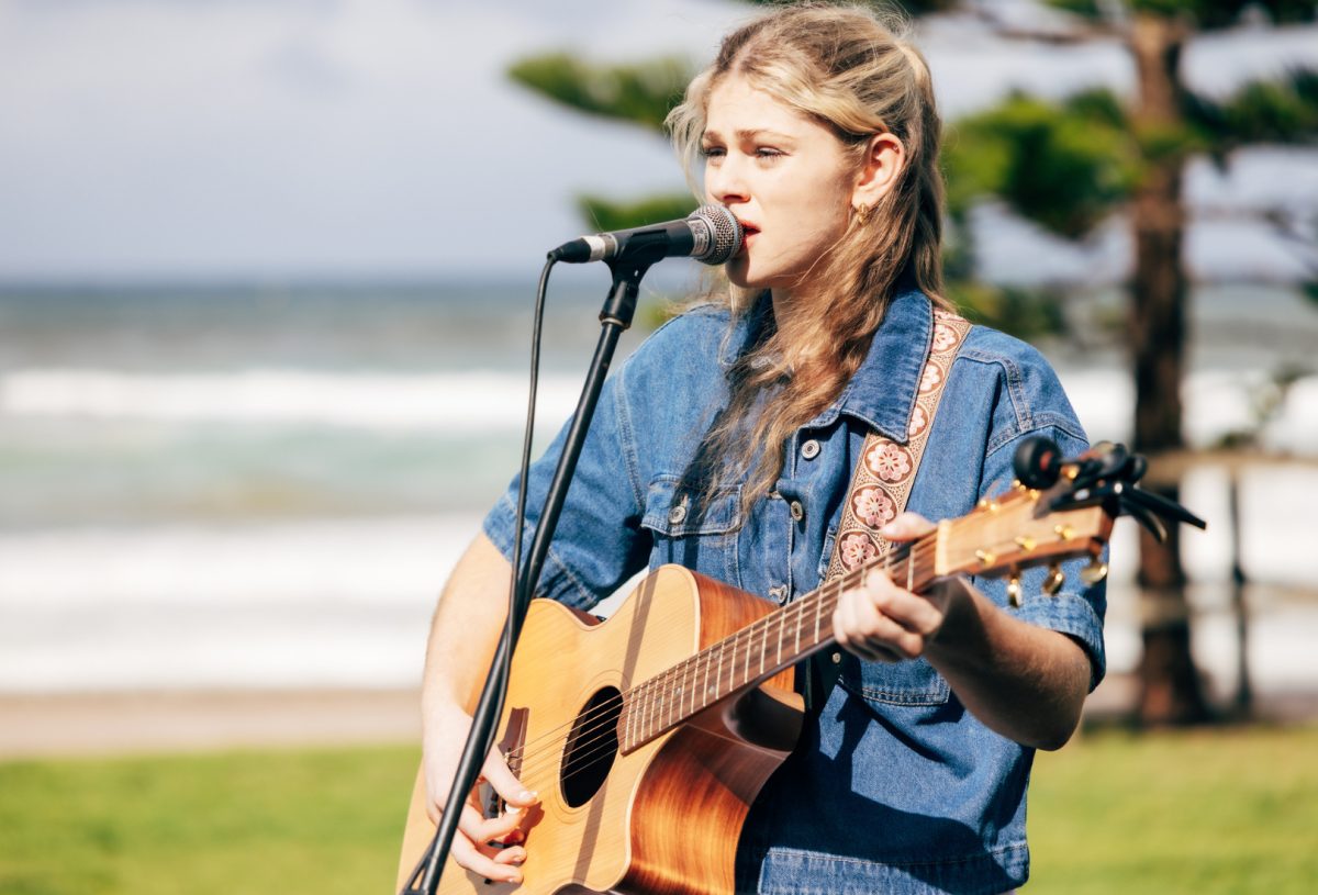 A young woman singing and playing guitar at a beachside park 