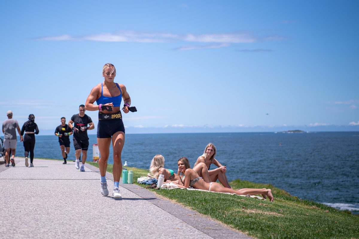 runners competing along a coastal path as sunbathing women look on