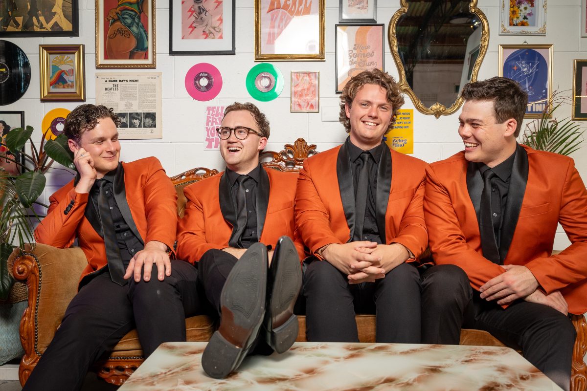 The singers of The 60 Four sit on a couch in orange jackets