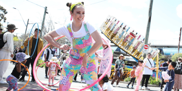 a person wearing colourful overalls hula-hooping in front of a carvnial ride