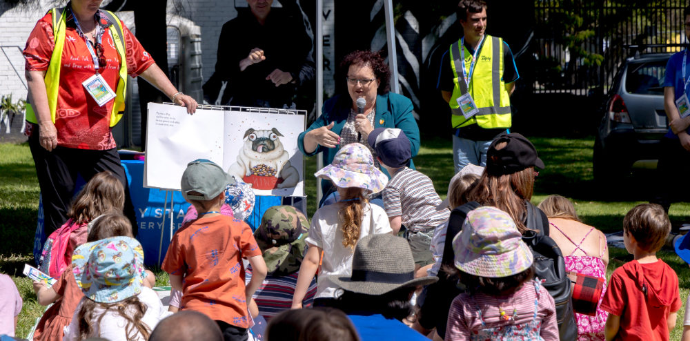 a group of children listening to a person reading a book outside