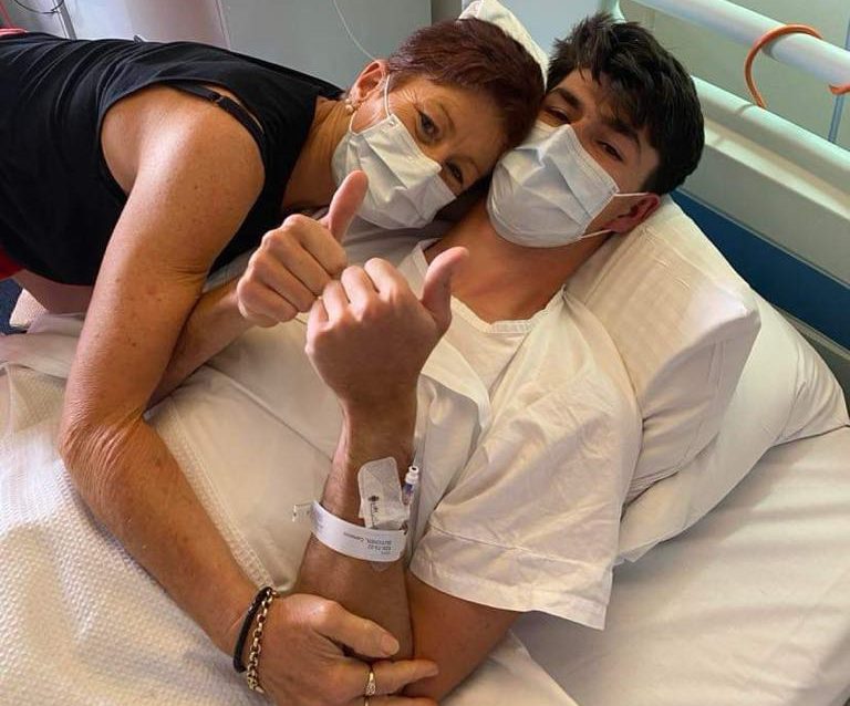 A young man in a hospital bed gives a thumbs up as his mum hugs him