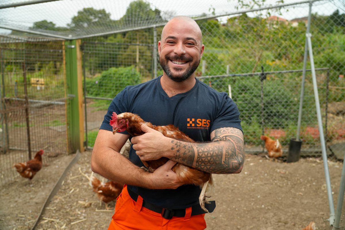 A man in an SES uniform stands in floodwaters holding a chicken and beaming at the camera.