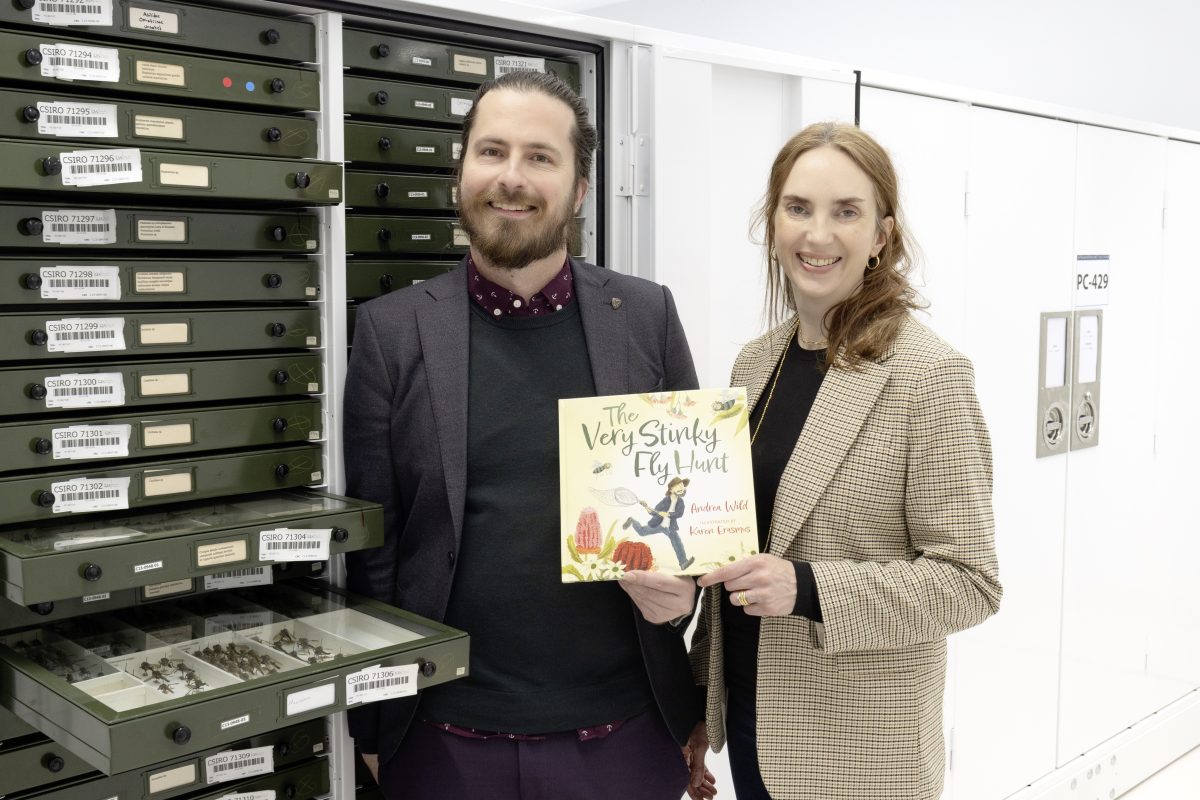 A man and a woman hold a child's picture book in front of a drawer full of dead fly specimens.