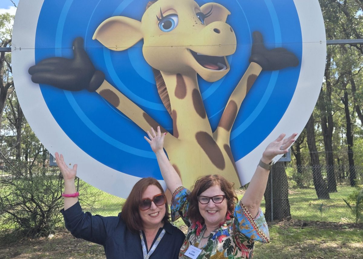 Development Manager Lisa Barber and Program Development Manager Michelle Weisbaum in front of an image of Harold the Giuraffe